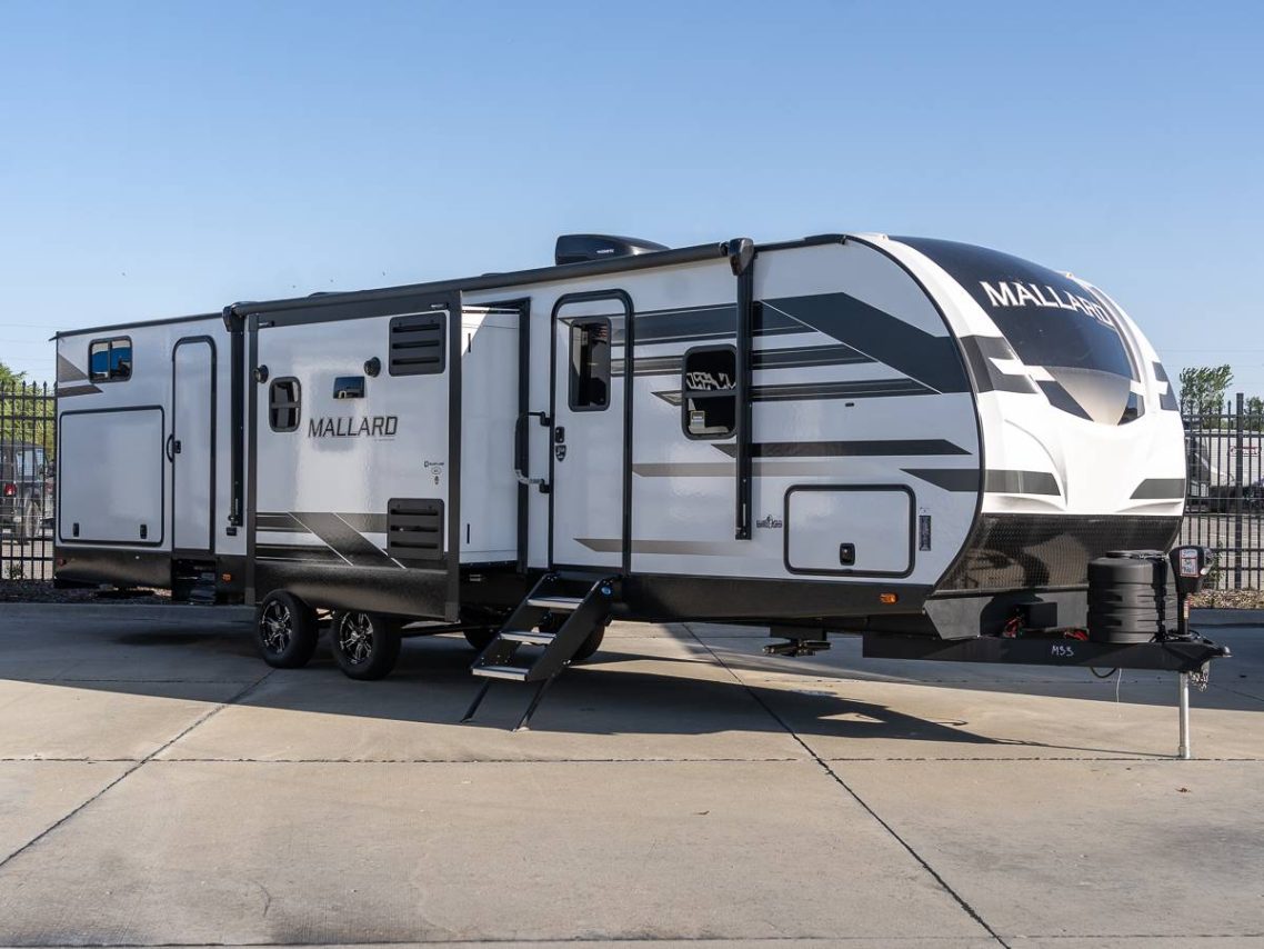 Heartland Mallard White and gray travel trailer with multiple doors and windows, parked outdoors.
